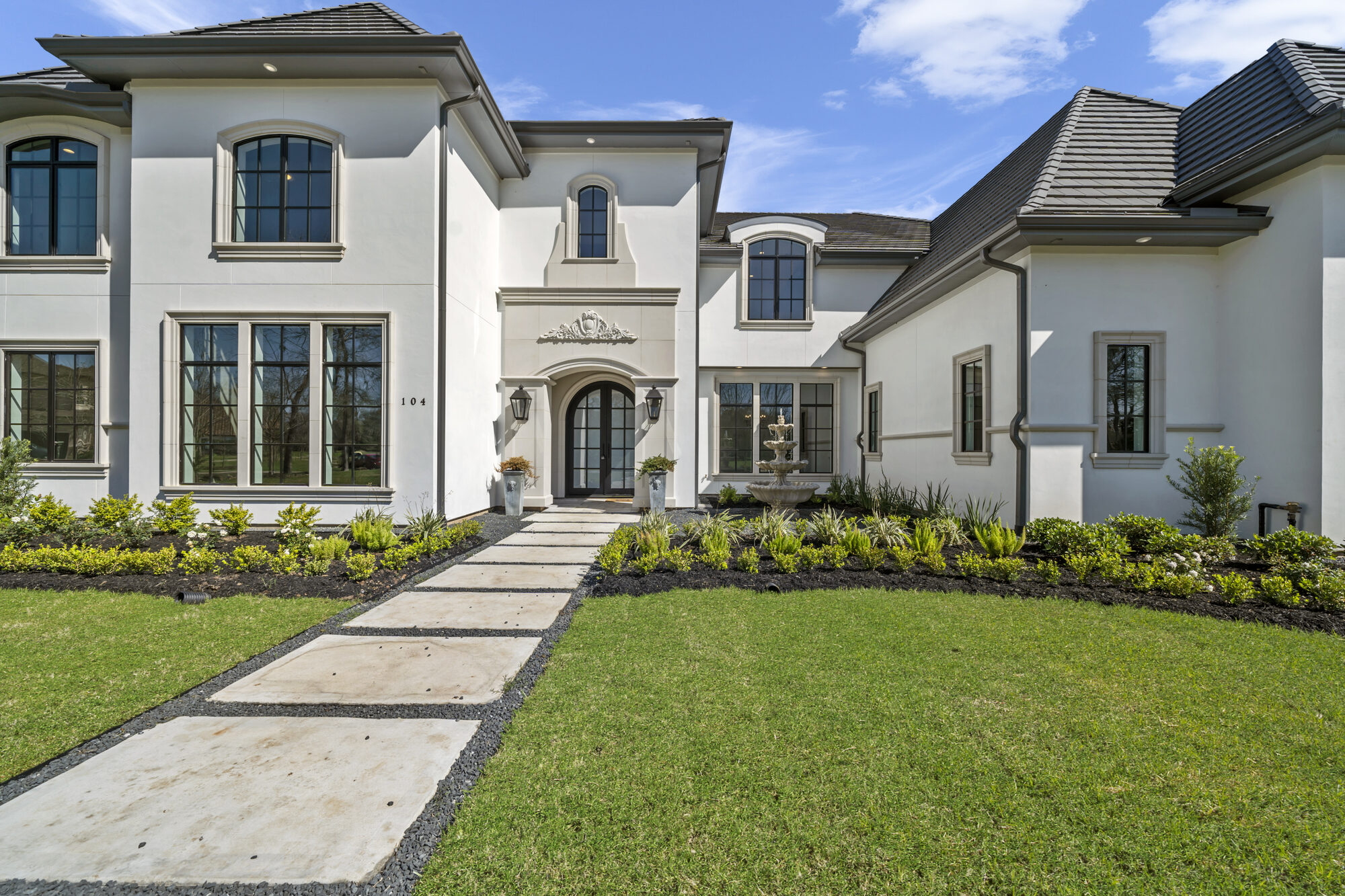 Front elevation of a white French-manor residence by Psalms Fine Homes, with an arched entry, stone fountain, and manicured lawn